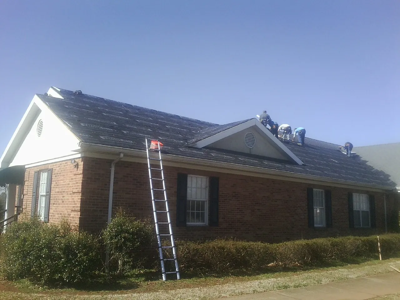 Crew tearing shingles off a brick ranch roof with a ladder set — Emergency Roof Repair work by our crew in Williamsfield