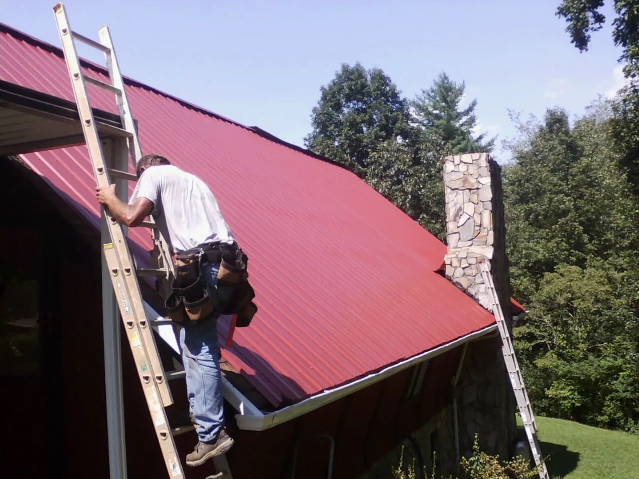Roofer on a ladder beside a red metal roof and stone chimney — Soffit & Fascia Repair by Roofing Brothers in Williamsfield